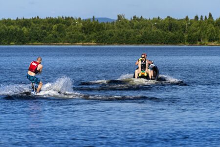 Finland, Lapland, Ivalo: Young man on modern jet ski water scooter has fun in the middle of the Finnish nature with lake water, cloud reflections, green trees, riverbank and blue sky. Jul 25, 2019のeditorial素材