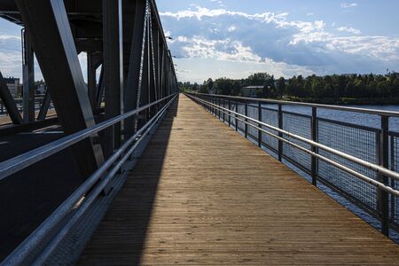 Republic of Finland, Rovaniemi: Pedestrian bridge as part of big railway bridge over Kemijoki river next to the city center of the Finnish town with sun, calm floating water, blue sky. Jul 26, 2019のeditorial素材