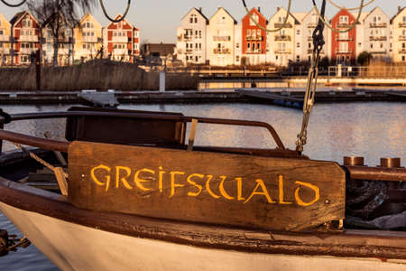 Germany, Greifswald, river Ryck - December 30, 2016: Wooden shield of an old sailing ship shows name of the northern German town Greifswald with colorful houses (Holzteichquartier) in the background.のeditorial素材