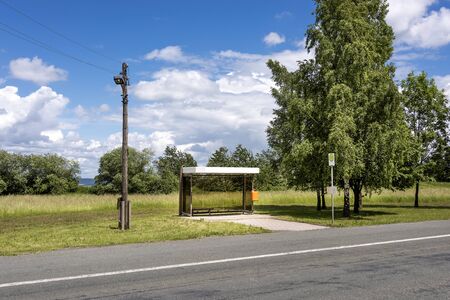 Germany, Thuringia, Bucha: Street scene with lonely bus stop, tarred road, lantern, green trees and cloudy blue sky - concept countryside, public transport, environmentの写真素材