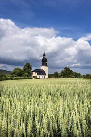 Germany, Thuringia, Unterwellenborn: Landscape panorama view with young corn field with small town village church, green trees and blue cloudy sky in the background - concept scenic agriculture homeの写真素材