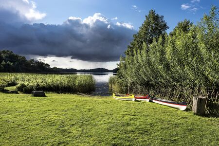 Germany, GroÃ Zecher, near Schaalsee: Dark dramatic thunderstorm clouds over natural area with meadow , lake, grass trees and colorful canoes.の写真素材