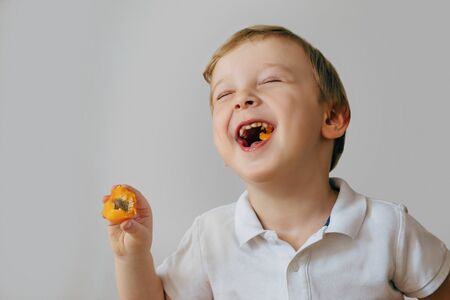 A three-year-old boy laughs after biting off a delicious orange fruit. The boy is wearing a white T-shirtの写真素材