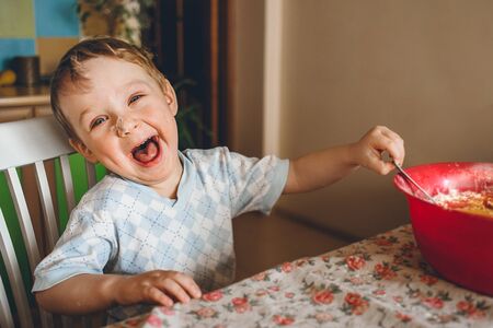 the little boy sits in the kitchen at the table and laughs at the camera. The child helps her mother to cook a pie. It stirs spoon the dough into a bowlの写真素材
