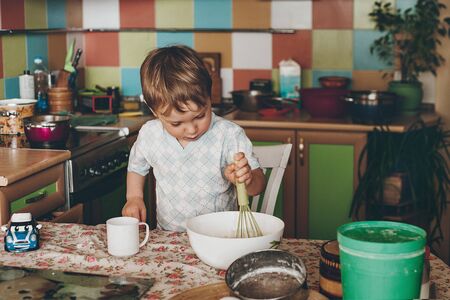 the little boy sits in the kitchen at the table. The child helps her mother to cook a pie. It stirs spoon the dough into a bowlの写真素材