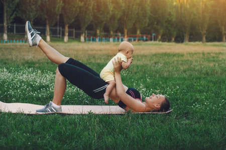 European sportive mother workout on nature grass lawn with toddler baby son in fitness clothing....