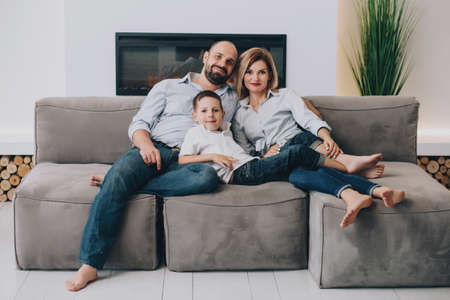 caucasian family is sitting on a gray sofa at home cuddling up and looking at the camera. Father...