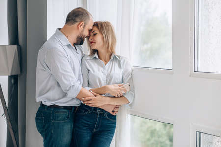 a woman and a man in jeans and shirts stand near a panoramic window and hug gently. Man embracing girlfriend at homeの写真素材