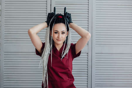 cheerful young woman with African braids on her head, in a red medical uniform and shows her horns with her hands on her head.の写真素材