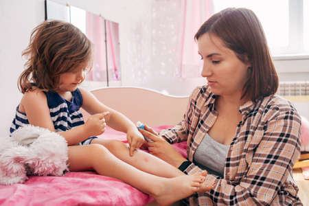 Baby girl sits on the bed in the bedroom and smears cream leg, next sits momの写真素材
