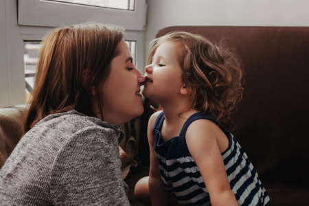 Little baby girl hugs and kisses her mother, daughter cuddles with mother at home near windowの写真素材