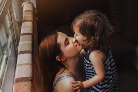 Little baby girl hugs and kisses her mother, daughter cuddles with mother at home near windowの写真素材