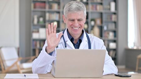 Cheerful Senior Doctor Doing Video Chat on Laptop while Sitting in Officeの写真素材