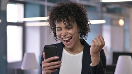 Portrait of Cheerful African Businesswoman Celebrating on Smartphone with Fistの写真素材
