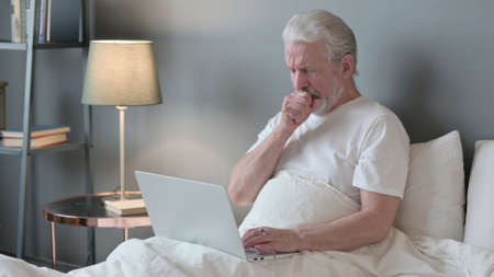 Senior Old Man with Laptop Coughing in Bedの写真素材