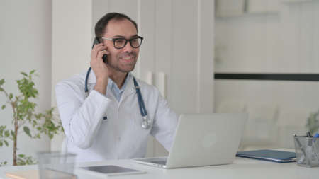 Middle Aged Doctor Talking on Smartphone while Working on Laptopの写真素材