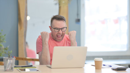 Young Adult Man Celebrating Online Success on Laptop in Officeの写真素材