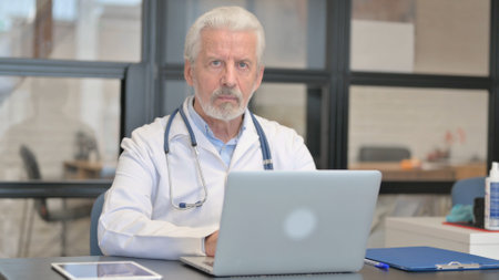 Senior Doctor Looking at Camera while Working on Laptopの写真素材