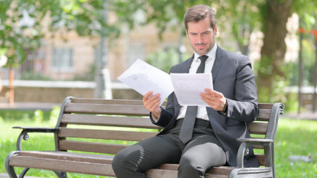 Middle Aged Businessman Reading Documents while Sitting Outdoor on Benchの写真素材