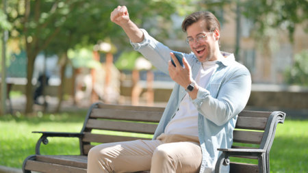 Casual Young Man Celebrating Success on Phone while Sitting in Parkの写真素材