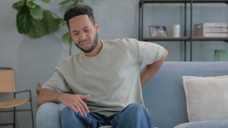 Hispanic Man with Back Pain Sitting on Sofa at Homeの写真素材
