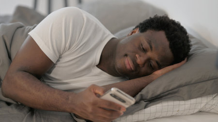 African American man using mobile phone while lying in bed at homeの写真素材