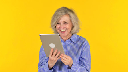 Senior woman using digital tablet on yellow background. Portrait of happy senior woman with tablet...