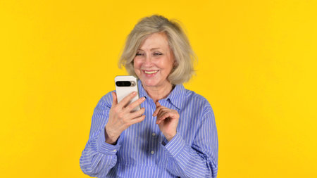 Old Woman Doing Video Chat via Phone on Yellow Backgroundの写真素材