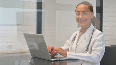 Multiracial Female Doctor Smiling at Camera while Working on Laptopの写真素材