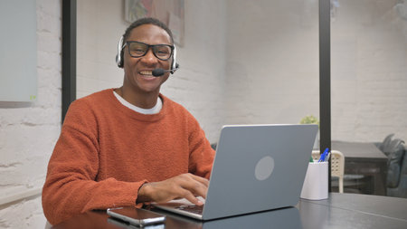 African American Man with Headset Smiling at Camera while Working on Laptop