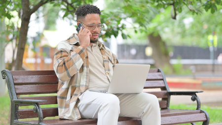 Hispanic Man Talking on Phone While Using Laptop Outdoorの写真素材