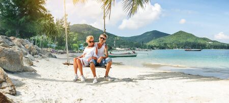mature couple rest on a swing at tropical beach, panoramaの写真素材