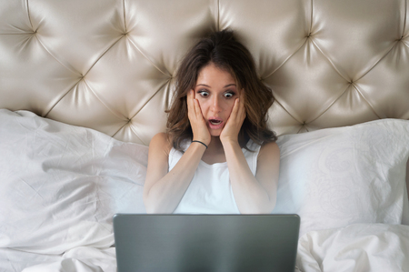 Woman looking in monitor with shock sitting in a bed in hotelの写真素材