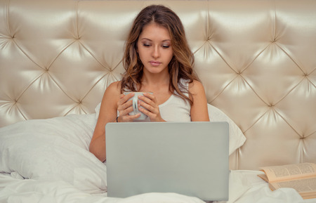 Woman sitting with laptop and a cup of coffee in a bedの写真素材