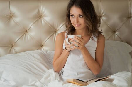 Cute girl sitting with a cup of coffee in a white bed in a hotelの写真素材