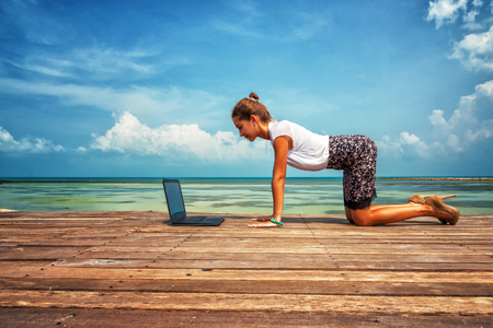 Woman in office suit doing yoga on the wooden floor with laptop. Young Lady has practice on the beach in front of sea view.の写真素材