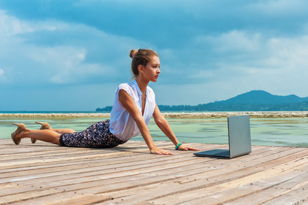 Woman in office suit doing yoga on the wooden floor with laptop. Young Lady has practice on the beach in front of sea view.の写真素材