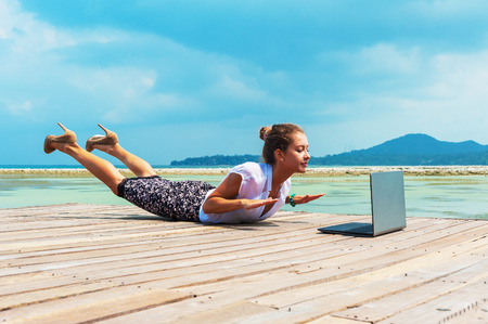Woman in office suit doing yoga on the wooden floor with laptop. Young Lady has practice on the beach in front of sea view.の写真素材