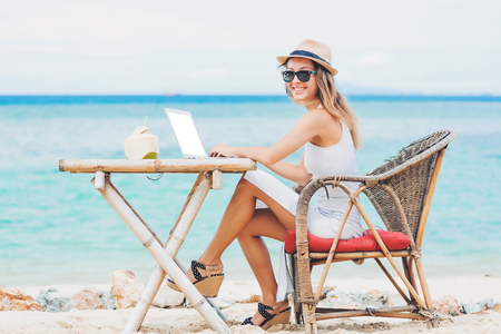 Young sexy woman with beautiful legs smiling on camera, using laptop with empty white screen on the beach. Girl Freelancer workingの写真素材