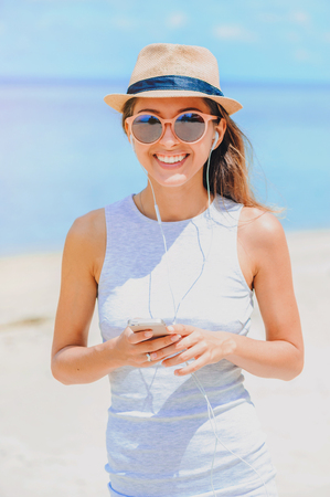 Young sexy woman in dress and sunglasses with hat on ocean background, listening music by phone on the beach. Girl with earphonesの写真素材