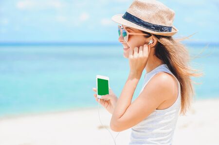Young sexy woman in dress, sunglasses and hat, holding phone with green screen on ocean background, listening music by phone on the beach. Girl with earphonesの写真素材