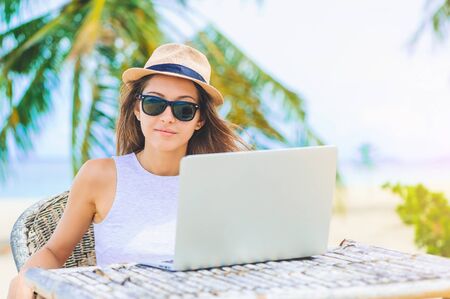 Young attractive woman in sunglasses and dress with hat working in laptop on the beach. Girl Freelancer workingの写真素材