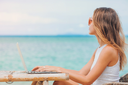 Young sexy woman freelancer in dress sitting at the table on ocean background, using laptop on the beach. Girl Freelancer workingの写真素材