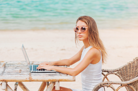 Young sexy woman freelancer in dress sitting at the table on ocean background, using laptop on the beach. Girl Freelancer workingの写真素材
