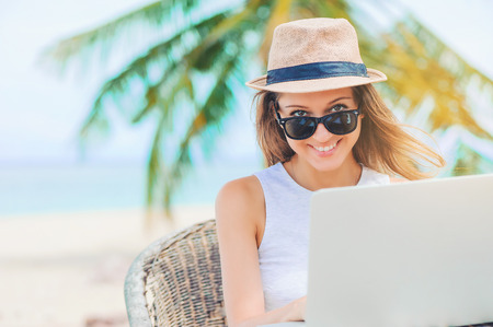 Young attractive woman in sunglasses and dress with hat working in laptop on the beach. Girl Freelancer workingの写真素材