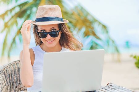 Young attractive woman in sunglasses and dress with hat working in laptop on the beach. Girl Freelancer workingの写真素材