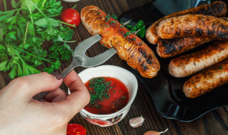 Woman's hands holding barbeque fork with grilled sausage on wooden table with sause, vegetables and sausagesの写真素材