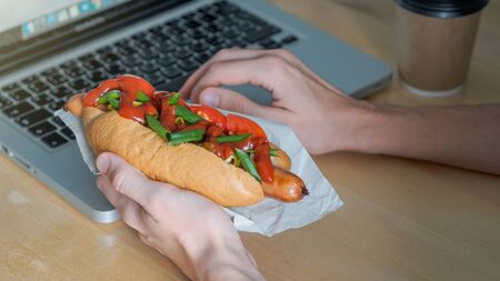 female freelancer hands holding hotdog and cup of coffee near the laptopの写真素材