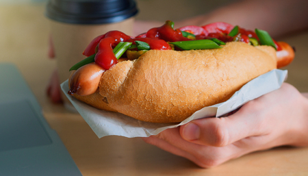 female freelancer hands holding hotdog and cup of coffee near the laptopの写真素材