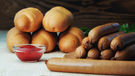 Ingredients for making homemade hot dogs. Sausages, fresh baked buns and ketchup sauce, served on wood chopping board over wooden background. Rustic styleの写真素材